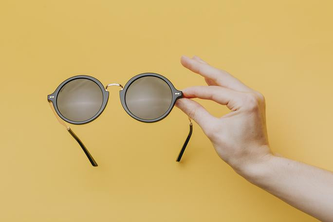 Hand holds a pair of sunglasses in front of a yellow background